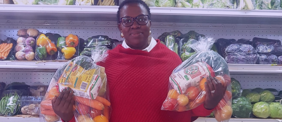 Gloria, founder of Maledi Fresh, standing in front of shelves of fresh produce while holding two bags of vegetables inside the Maledi Fresh store.