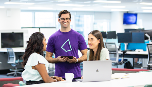 A man in an Iress branded t-shirt smiles at the camera while two female colleagues have a discussion in the foreground